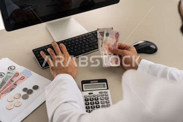 استخدام الحاسبة الرقمية في الأعمال الاقتصادية، محاولة إدارة المصروف المنزلي بتدبر، السعي نحو التوفير والادخار، صورة مقربة من الخلف لرجل عربي خليجي اماراتي يرتدي الكندورة والغترة البيضاء يجلس على كرسي مريح يقوم بالحسابات الشهرية المالية