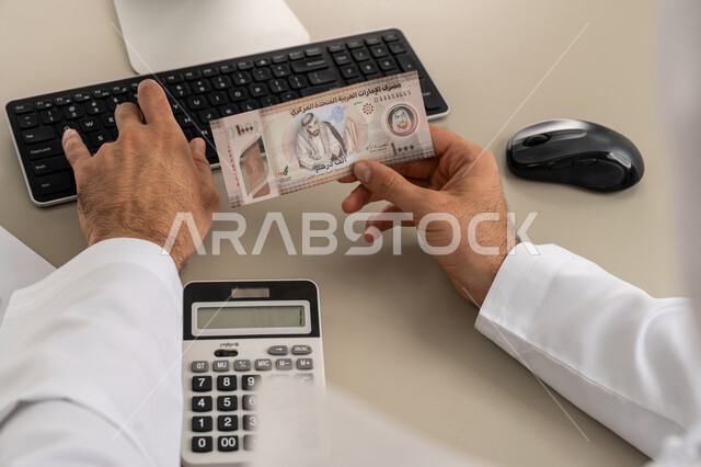 Trying to manage household expenses wisely, striving towards saving and saving, using a digital calculator in economic activities, a close-up image from the back of an Arab Gulf Emirati man wearing a white kandura and ghutra sitting on a comfortable chair doing monthly financial calculations