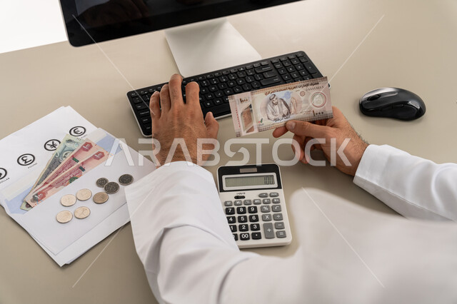 محاولة إدارة المصروف المنزلي بتدبر، السعي نحو التوفير والادخار، استخدام الحاسبة الرقمية في الأعمال الاقتصادية، صورة مقربة من الخلف لرجل عربي خليجي اماراتي يرتدي الكندورة والغترة البيضاء يجلس على كرسي مريح يقوم بالحسابات الشهرية المالية