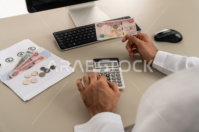 استخدام الحاسبة الرقمية في الأعمال الاقتصادية، محاولة إدارة المصروف المنزلي بتدبر، السعي نحو التوفير والادخار، صورة مقربة من الخلف لرجل عربي خليجي اماراتي يرتدي الكندورة والغترة البيضاء يجلس على كرسي مريح يقوم بالحسابات الشهرية المالية
