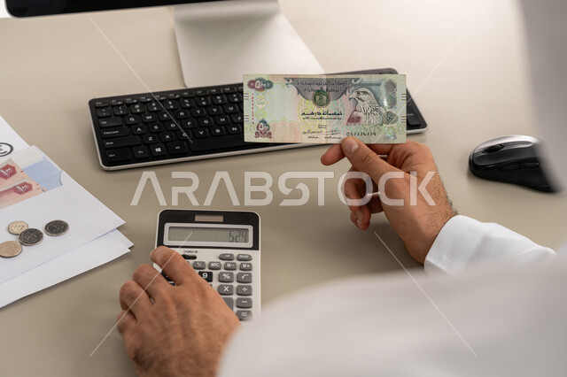 محاولة إدارة المصروف المنزلي بتدبر، السعي نحو التوفير والادخار، استخدام الحاسبة الرقمية في الأعمال الاقتصادية، صورة مقربة من الخلف لرجل عربي خليجي اماراتي يرتدي الكندورة والغترة البيضاء يجلس على كرسي مريح يقوم بالحسابات الشهرية المالية