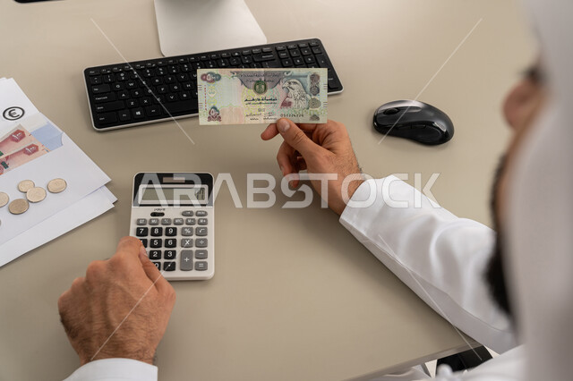 استخدام الحاسبة الرقمية في الأعمال الاقتصادية، محاولة إدارة المصروف المنزلي بتدبر، السعي نحو التوفير والادخار، صورة مقربة من الخلف لرجل عربي خليجي اماراتي يرتدي الكندورة والغترة البيضاء يجلس على كرسي مريح يقوم بالحسابات الشهرية المالية