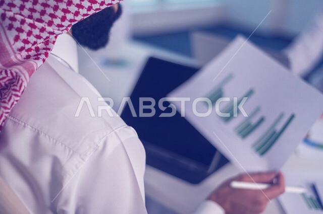 رجل اعمال عربي خليجي سعودي يجلس امام الحاسوب المحمول ، يحمل بيده ورقة المخطط المالي ، اجتماع فريق العمل ، مناقشة الرسم البياني المالي ، خطة الاستثمار ، استشارات مالية ومحاسبية، تحقيق اهداف العمل