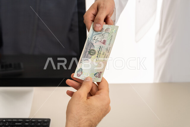 تسجيل تسديدات العملاء على جهاز الحاسوب المحمول، العملة المتداولة في أسواق الامارات العربية المتحدة،  بورتريه لرجل عربي خليجي اماراتي يرتدي الكندورة والغترة البيضاء يقوم بدفع الفواتير وحسابات المشتريات، استخدام الاجهزة التقنية الحديثة والمتطورة، خلفية بيضاء