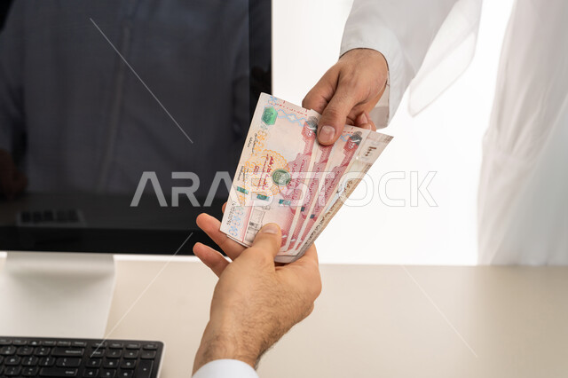 تسجيل تسديدات العملاء على جهاز الحاسوب المحمول، العملة المتداولة في أسواق الامارات العربية المتحدة،  بورتريه لرجل عربي خليجي اماراتي يرتدي الكندورة والغترة البيضاء يقوم بدفع الفواتير وحسابات المشتريات، استخدام الاجهزة التقنية الحديثة والمتطورة، خلفية بيضاء