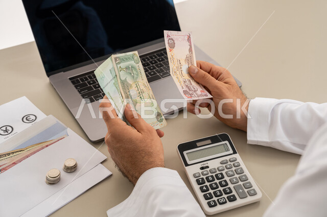محاولة إدارة المصروف المنزلي بتدبر، السعي نحو التوفير والادخار، صورة مقربة من الخلف لرجل عربي خليجي اماراتي يرتدي الكندورة والغترة البيضاء يجلس على كرسي مريح يقوم بالحسابات الشهرية المالية، استخدام الحاسبة الرقمية في الأعمال الاقتصادية
