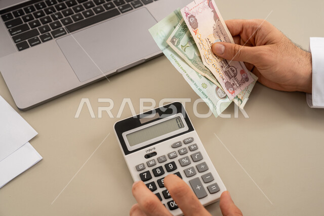 استخدام الحاسبة الرقمية في الأعمال الاقتصادية، محاولة إدارة المصروف المنزلي بتدبر، السعي نحو التوفير والادخار، صورة مقربة من الخلف لرجل عربي خليجي اماراتي يرتدي الكندورة والغترة البيضاء يجلس على كرسي مريح يقوم بالحسابات الشهرية المالية