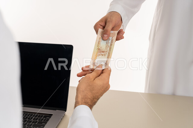 تسجيل تسديدات العملاء على جهاز الحاسوب المحمول، العملة المتداولة في أسواق الامارات العربية المتحدة،  بورتريه لرجل عربي خليجي اماراتي يرتدي الكندورة والغترة البيضاء يقوم بدفع الفواتير وحسابات المشتريات، استخدام الاجهزة التقنية الحديثة والمتطورة، خلفية بيضاء