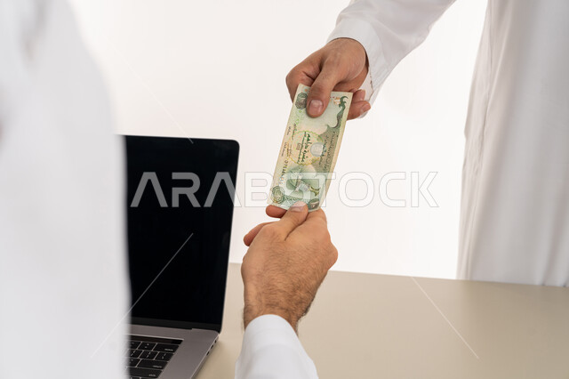 تسجيل تسديدات العملاء على جهاز الحاسوب المحمول، العملة المتداولة في أسواق الامارات العربية المتحدة،  بورتريه لرجل عربي خليجي اماراتي يرتدي الكندورة والغترة البيضاء يقوم بدفع الفواتير وحسابات المشتريات، استخدام الاجهزة التقنية الحديثة والمتطورة، خلفية بيضاء