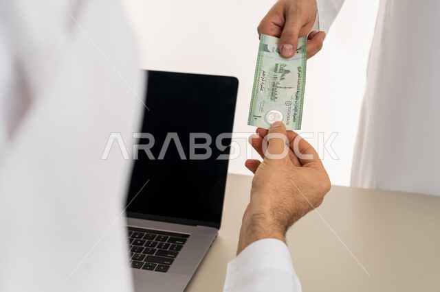 تسجيل تسديدات العملاء على جهاز الحاسوب المحمول، العملة المتداولة في أسواق الامارات العربية المتحدة،  بورتريه لرجل عربي خليجي اماراتي يرتدي الكندورة والغترة البيضاء يقوم بدفع الفواتير وحسابات المشتريات، استخدام الاجهزة التقنية الحديثة والمتطورة، خلفية بيضاء