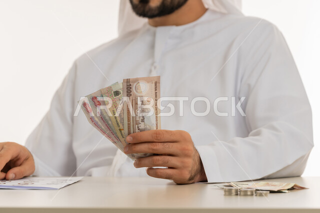 عرض الإصدار الجديد للنقود الاماراتية، العملة المتداولة في أسواق الامارات العربية المتحدة،  بورتريه لرجل عربي خليجي اماراتي يرتدي الكندورة والغترة يمسك بيده الفلوس، الأعمال الاقتصادية المالية، محاولة إدارة المصروف المنزلي بتدبر، السعي نحو الادخار، خلفية بيضاء