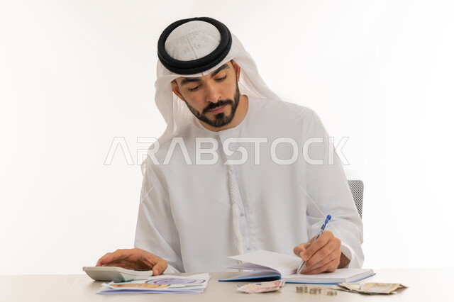 محاولة إدارة المصروف المنزلي بتدبر، السعي نحو التوفير والادخار، استخدام الحاسبة الرقمية في الأعمال الاقتصادية، بورتريه لرجل عربي خليجي اماراتي يرتدي الكندورة والغترة البيضاء يجلس على كرسي مريح يقوم بالحسابات الشهرية المالية، خلفية بيضاء