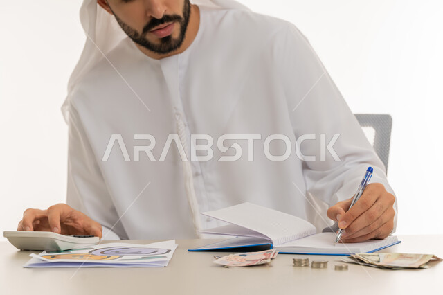 محاولة إدارة المصروف المنزلي بتدبر، السعي نحو التوفير والادخار، استخدام الحاسبة الرقمية في الأعمال الاقتصادية، بورتريه لرجل عربي خليجي اماراتي يرتدي الكندورة والغترة البيضاء يجلس على كرسي مريح يقوم بالحسابات الشهرية المالية، خلفية بيضاء