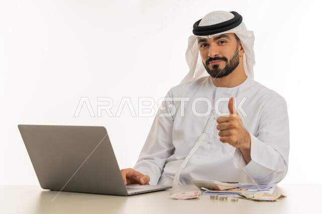 Success and achievement of goals, mark of quality and excellence, striving for savings and thrift, portrait of an Arab Gulf Emirati man wearing a kandura and ghutra raising his thumb up with gestures of acceptance, doing monthly calculations on a laptop, admiration and satisfaction for achieving a certain thing, white background