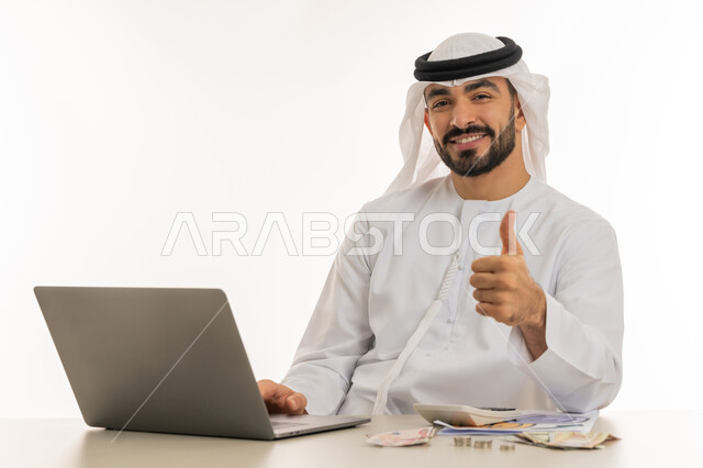 النجاح وتحقيق الأهداف، علامة الجودة والامتياز، السعي نحو التوفير والادخار، بورتريه لرجل عربي خليجي اماراتي يرتدي الكندورة والغترة يرفع اصبع الابهام للاعلى بإيماءات القبول يقوم بالحسابات الشهرية بواسطة الحاسوب المحمول، الاعجاب والرضا لتحقيق أمر معين، خلفية بيضاء