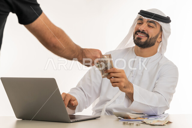 Recording customer payments on laptop, currency in UAE markets, portrait of an Emirati Gulf Arab man wearing a white kandura and ghutra paying bills and purchase accounts, using modern and advanced technical devices, white background