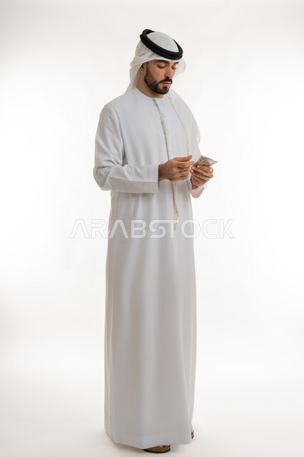 ادخار النقود لوقت الحاجة، العملة المتداولة في أسواق الامارات العربية المتحدة، القيام بالحسابات الشهرية، محاولة الاقتصاد في المصروف المنزلي، بورتريه لرجل عربي خليجي اماراتي يرتدي الكندورة والغترة يقوم بعد النقود والأوراق المالية بإيماءات التركيز، خلفية بيضاء
