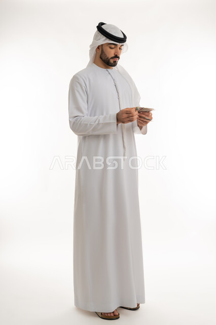 ادخار النقود لوقت الحاجة، العملة المتداولة في أسواق الامارات العربية المتحدة، القيام بالحسابات الشهرية، محاولة الاقتصاد في المصروف المنزلي، بورتريه لرجل عربي خليجي اماراتي يرتدي الكندورة والغترة يقوم بعد النقود والأوراق المالية بإيماءات التركيز، خلفية بيضاء