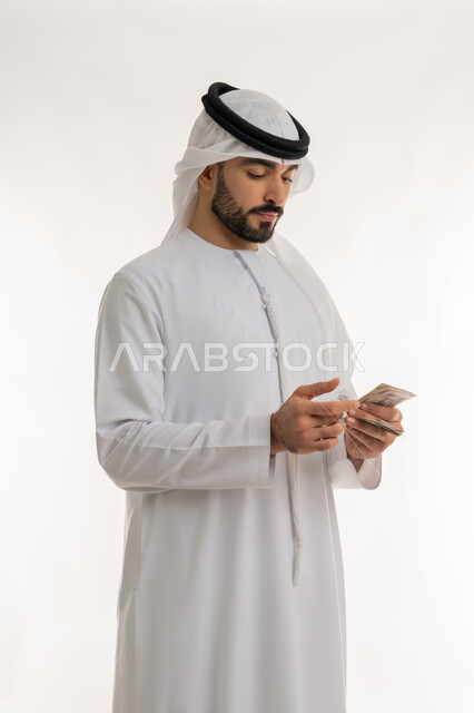 ادخار النقود لوقت الحاجة، العملة المتداولة في أسواق الامارات العربية المتحدة، القيام بالحسابات الشهرية، محاولة الاقتصاد في المصروف المنزلي، بورتريه لرجل عربي خليجي اماراتي يرتدي الكندورة والغترة يقوم بعد النقود والأوراق المالية بإيماءات التركيز، خلفية بيضاء