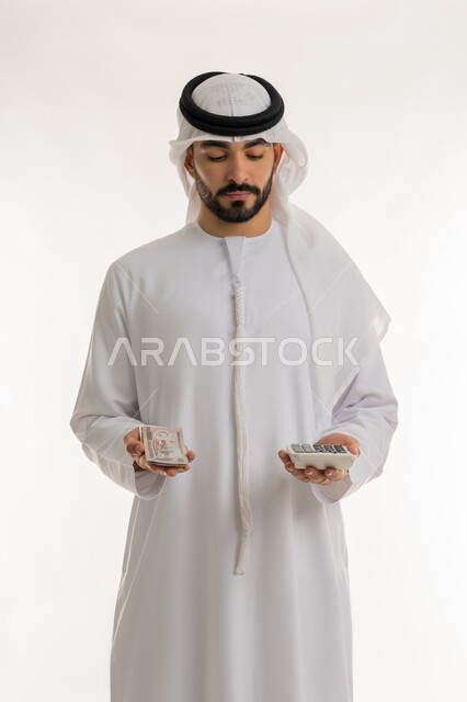 الأعمال الاقتصادية المالية،  محاولة إدارة المصروف المنزلي بتدبر، بورتريه لرجل عربي خليجي اماراتي يرتدي الكندورة والغترة يمسك بإحدى يديه الحاسبة الرقمية وباليد الأخرى مجموعة من النقود، السعي نحو التوفير والادخار، خلفية بيضاء