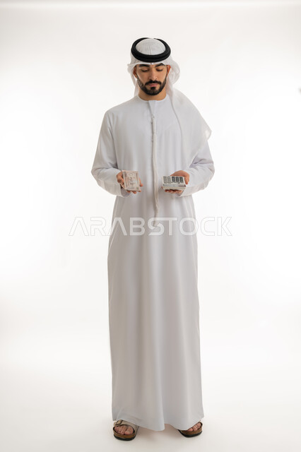 الأعمال الاقتصادية المالية،  محاولة إدارة المصروف المنزلي بتدبر، بورتريه لرجل عربي خليجي اماراتي يرتدي الكندورة والغترة يمسك بإحدى يديه الحاسبة الرقمية وباليد الأخرى مجموعة من النقود، السعي نحو التوفير والادخار، خلفية بيضاء