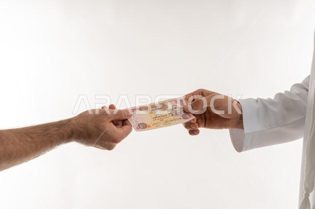 مفهوم الدفع نقدا، عملية تسديد النقود بواسطة الدراهم، استلام وتسليم الأمانات، العملة المتداولة في أسواق الامارات العربية المتحدة، الأعمال الاقتصادية المالية، بورتريه مقرب لرجل عربي خليجي اماراتي يرتدي الكندورة يقوم بدفع الفواتير وحسابات المشتريات، خلفية بيضاء