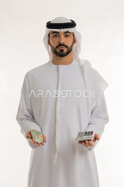 Looking at the camera with gestures of self-confidence, financial economics, trying to manage household expenses wisely, portrait of an Arab Gulf Emirati man wearing a kandura and a ghutra holding a digital calculator in one hand and a bunch of money in the other, striving towards saving and saving, white background