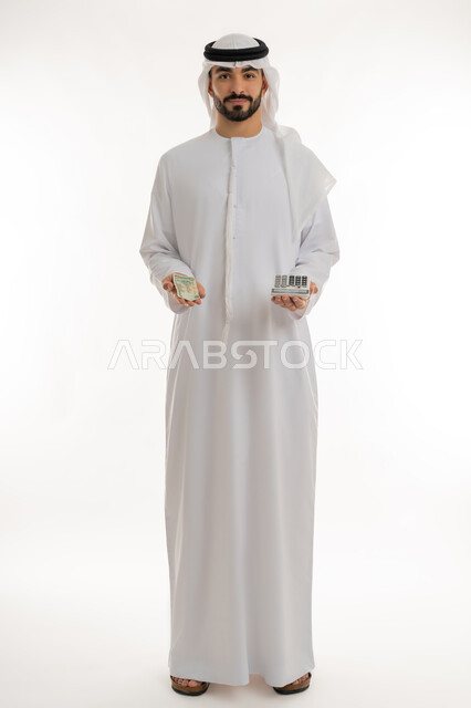 النظر الى الكاميرا بايماءات الثقة بالنفس ، الأعمال الاقتصادية المالية، محاولة إدارة المصروف المنزلي بتدبر، بورتريه لرجل عربي خليجي اماراتي يرتدي الكندورة والغترة يمسك بإحدى يديه الحاسبة الرقمية وباليد الأخرى مجموعة من النقود، السعي نحو التوفير والادخار، خلفية بيضاء