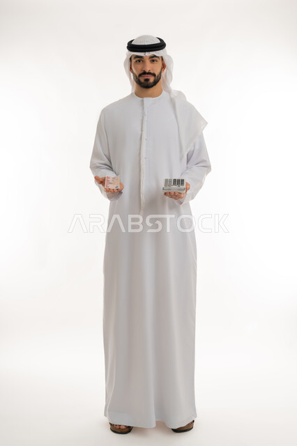 النظر الى الكاميرا بايماءات الثقة بالنفس ، الأعمال الاقتصادية المالية، محاولة إدارة المصروف المنزلي بتدبر، بورتريه لرجل عربي خليجي اماراتي يرتدي الكندورة والغترة يمسك بإحدى يديه الحاسبة الرقمية وباليد الأخرى مجموعة من النقود، السعي نحو التوفير والادخار، خلفية بيضاء