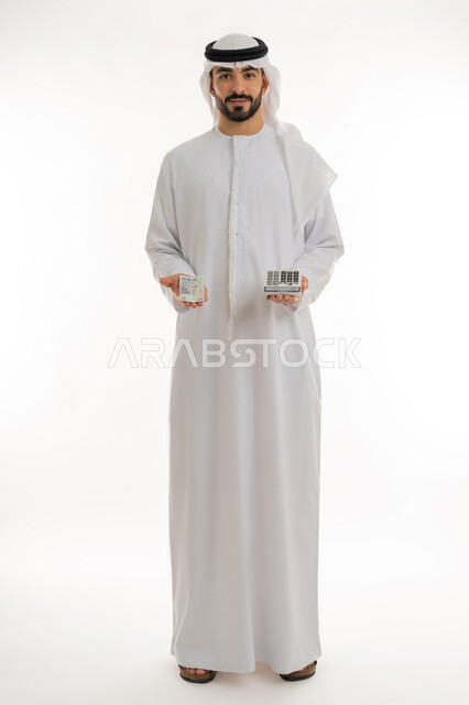 النظر الى الكاميرا بايماءات الثقة بالنفس ، الأعمال الاقتصادية المالية، محاولة إدارة المصروف المنزلي بتدبر، بورتريه لرجل عربي خليجي اماراتي يرتدي الكندورة والغترة يمسك بإحدى يديه الحاسبة الرقمية وباليد الأخرى مجموعة من النقود، السعي نحو التوفير والادخار، خلفية بيضاء