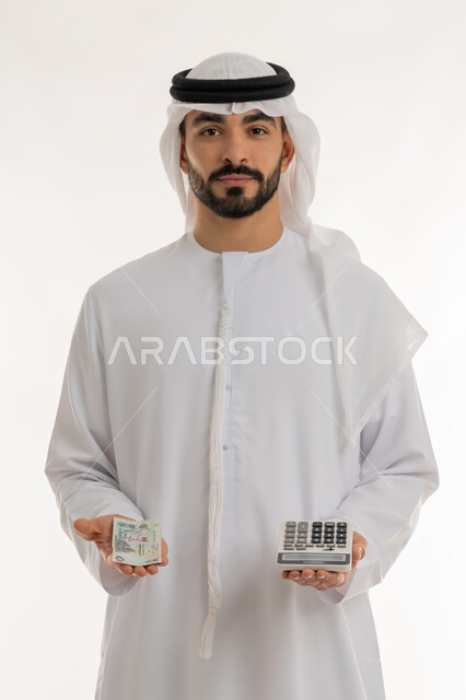 النظر الى الكاميرا بايماءات الثقة بالنفس ، الأعمال الاقتصادية المالية، محاولة إدارة المصروف المنزلي بتدبر، بورتريه لرجل عربي خليجي اماراتي يرتدي الكندورة والغترة يمسك بإحدى يديه الحاسبة الرقمية وباليد الأخرى مجموعة من النقود، السعي نحو التوفير والادخار، خلفية بيضاء