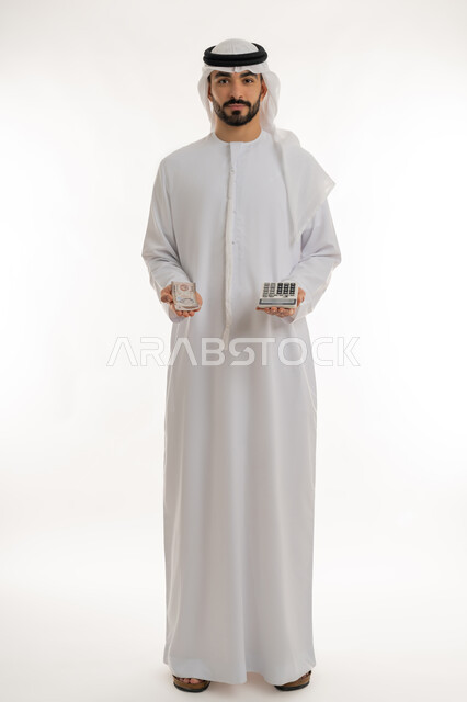 النظر الى الكاميرا بايماءات الثقة بالنفس ، الأعمال الاقتصادية المالية، محاولة إدارة المصروف المنزلي بتدبر، بورتريه لرجل عربي خليجي اماراتي يرتدي الكندورة والغترة يمسك بإحدى يديه الحاسبة الرقمية وباليد الأخرى مجموعة من النقود، السعي نحو التوفير والادخار، خلفية بيضاء