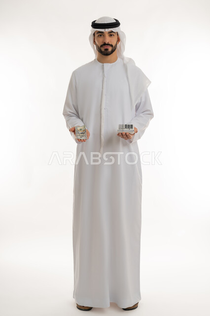 النظر الى الكاميرا بايماءات الثقة بالنفس ، الأعمال الاقتصادية المالية، محاولة إدارة المصروف المنزلي بتدبر، بورتريه لرجل عربي خليجي اماراتي يرتدي الكندورة والغترة يمسك بإحدى يديه الحاسبة الرقمية وباليد الأخرى مجموعة من النقود، السعي نحو التوفير والادخار، خلفية بيضاء