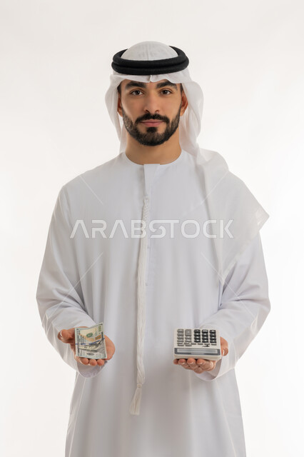 النظر الى الكاميرا بايماءات الثقة بالنفس ، الأعمال الاقتصادية المالية، محاولة إدارة المصروف المنزلي بتدبر، بورتريه لرجل عربي خليجي اماراتي يرتدي الكندورة والغترة يمسك بإحدى يديه الحاسبة الرقمية وباليد الأخرى مجموعة من النقود، السعي نحو التوفير والادخار، خلفية بيضاء