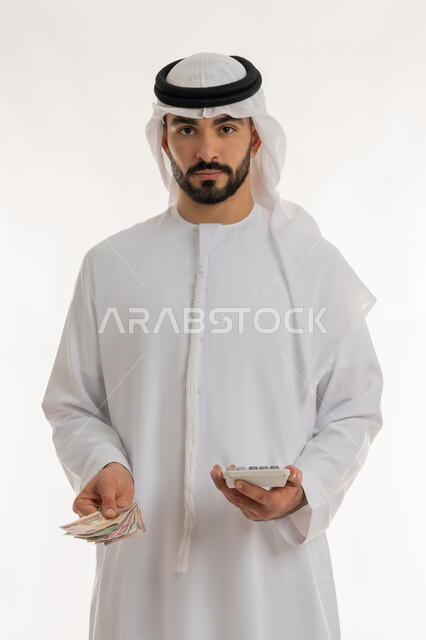 النظر الى الكاميرا بايماءات الثقة بالنفس ، الأعمال الاقتصادية المالية، محاولة إدارة المصروف المنزلي بتدبر، بورتريه لرجل عربي خليجي اماراتي يرتدي الكندورة والغترة يمسك بإحدى يديه الحاسبة الرقمية وباليد الأخرى مجموعة من النقود، السعي نحو التوفير والادخار، خلفية بيضاء