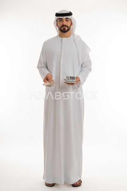 النظر الى الكاميرا بايماءات الثقة بالنفس ، الأعمال الاقتصادية المالية، محاولة إدارة المصروف المنزلي بتدبر، بورتريه لرجل عربي خليجي اماراتي يرتدي الكندورة والغترة يمسك بإحدى يديه الحاسبة الرقمية وباليد الأخرى مجموعة من النقود، السعي نحو التوفير والادخار، خلفية بيضاء