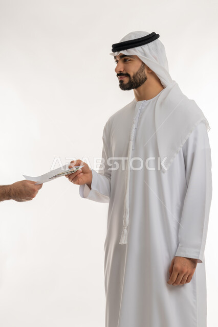 مفهوم الدفع نقدا، عملية تسديد النقود بواسطة الدراهم، استلام وتسليم الأمانات، العملة المتداولة في أسواق الامارات العربية المتحدة، الأعمال الاقتصادية المالية، بورتريه مقرب لرجل عربي خليجي اماراتي يرتدي الكندورة يقوم بدفع الفواتير وحسابات المشتريات، خلفية بيضاء