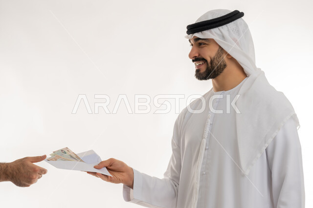 مفهوم الدفع نقدا، عملية تسديد النقود بواسطة الدراهم، استلام وتسليم الأمانات، العملة المتداولة في أسواق الامارات العربية المتحدة، الأعمال الاقتصادية المالية، بورتريه مقرب لرجل عربي خليجي اماراتي يرتدي الكندورة يقوم بدفع الفواتير وحسابات المشتريات، خلفية بيضاء