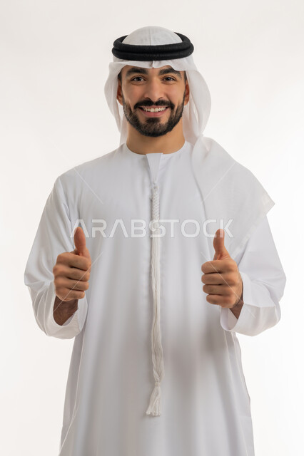 النظر الى الكاميرا بتعابير السعادة والسرور، النجاح وتحقيق الأهداف، علامة الجودة والامتياز, بورتريه مقرب لرجل عربي خليجي اماراتي مبتسم يرتدي الكندورة والغترة البيضاء يرفع اصبع الابهام للاعلى بإيماءات الموافقة والقبول, الاعجاب والرضا لتحقيق أمر معين, خلفية بيضاء