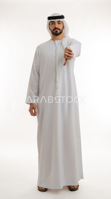 مفهوم التقييم السلبي والانزعاج، القلق والفشل في تحقيق أمر ما، بورتريه لرجل عربي خليجي إماراتي يرتدي الكندورة والغترة ينظر الى الكاميرا يشير بإصبع الابهام الى الاسفل بإيماءات الانزعاج،  تعابير الرفض وعدم القبول والرضا، الجسم بالطول الكامل، خلفية بيضاء