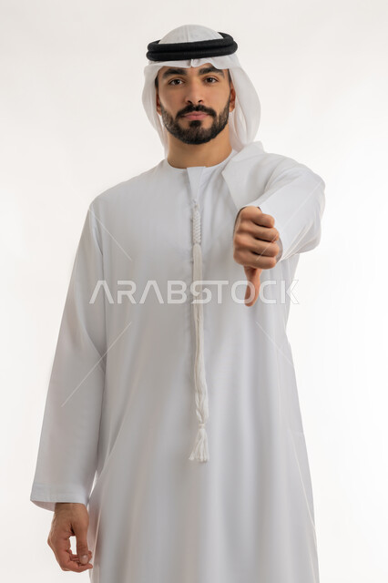 القلق والفشل في تحقيق أمر ما، مفهوم التقييم السلبي والانزعاج، بورتريه لرجل عربي خليجي إماراتي يرتدي الكندورة والغترة ينظر الى الكاميرا يشير بإصبع الابهام الى الاسفل بإيماءات الانزعاج،  تعابير الرفض وعدم القبول والرضا، خلفية بيضاء