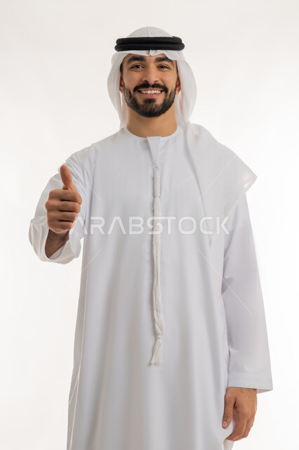 النظر الى الكاميرا بتعابير السعادة والسرور، النجاح وتحقيق الأهداف، علامة الجودة والامتياز, بورتريه مقرب لرجل عربي خليجي اماراتي مبتسم يرتدي الكندورة والغترة البيضاء يرفع اصبع الابهام للاعلى بإيماءات الموافقة والقبول, الاعجاب والرضا لتحقيق أمر معين, خلفية بيضاء