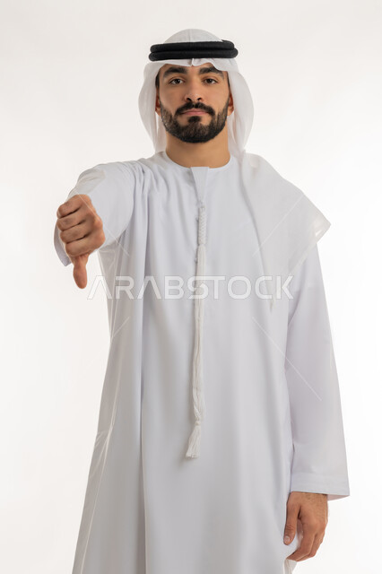 القلق والفشل في تحقيق أمر ما، مفهوم التقييم السلبي والانزعاج، بورتريه لرجل عربي خليجي إماراتي يرتدي الكندورة والغترة ينظر الى الكاميرا يشير بإصبع الابهام الى الاسفل بإيماءات الانزعاج،  تعابير الرفض وعدم القبول والرضا، خلفية بيضاء