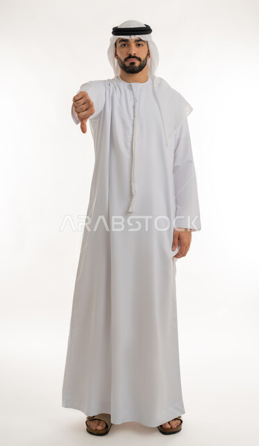مفهوم التقييم السلبي والانزعاج، القلق والفشل في تحقيق أمر ما، بورتريه لرجل عربي خليجي إماراتي يرتدي الكندورة والغترة ينظر الى الكاميرا يشير بإصبع الابهام الى الاسفل بإيماءات الانزعاج،  تعابير الرفض وعدم القبول والرضا، الجسم بالطول الكامل، خلفية بيضاء