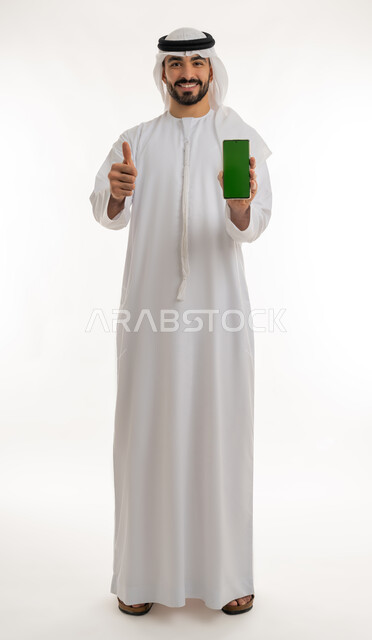 الاعجاب والرضا لتحقيق أمر معين, عرض شاشة خضراء عبر الهاتف المحمول، النجاح وتحقيق الأهداف، علامة الجودة والامتياز, بورتريه مقرب لرجل عربي خليجي اماراتي مبتسم يرتدي الكندورة والغترة البيضاء يرفع اصبع الابهام للاعلى بإيماءات الموافقة والقبول, خلفية بيضاء