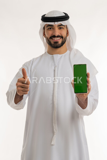 الاعجاب والرضا لتحقيق أمر معين, عرض شاشة خضراء عبر الهاتف المحمول، النجاح وتحقيق الأهداف، علامة الجودة والامتياز, بورتريه مقرب لرجل عربي خليجي اماراتي مبتسم يرتدي الكندورة والغترة البيضاء يرفع اصبع الابهام للاعلى بإيماءات الموافقة والقبول, خلفية بيضاء