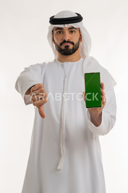 عرض شاشة خضراء عبر الهاتف المحمول، الفشل في تحقيق أمر ما، مفهوم التقييم السلبي والانزعاج، بورتريه لرجل عربي خليجي إماراتي يرتدي الكندورة والغترة ينظر الى الكاميرا يشير بإصبع الابهام الى الاسفل بإيماءات الانزعاج،  تعابير الرفض وعدم القبول، خلفية بيضاء، كروما