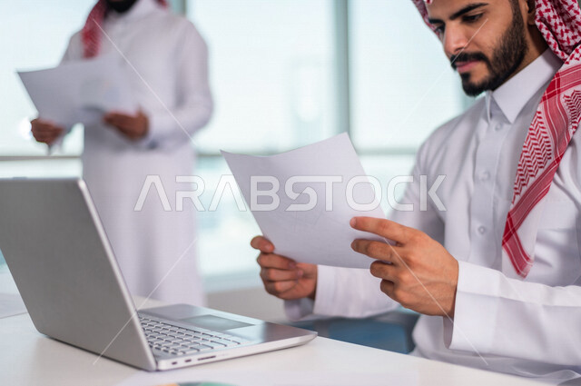 رجل اعمال عربي خليجي سعودي يجلس امام الحاسوب المحمول ، يحمل بيده ورقة المخطط المالي ، اجتماع فريق العمل ، مناقشة الرسم البياني المالي ، خطة الاستثمار ، استشارات مالية ومحاسبية، تحقيق اهداف العمل