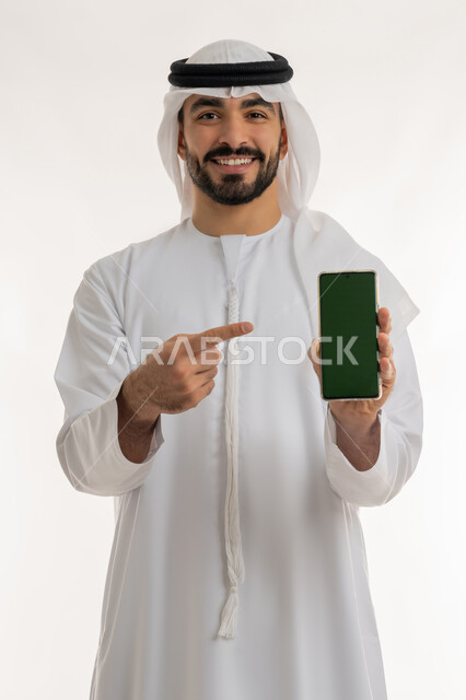 Using advanced technology for digital marketing, displaying mobile applications and services, portrait of a young Emirati Gulf Arab man wearing a kandura and a ghutra pointing his index finger to a mobile device with a blank green screen, looking at the camera with gestures of happiness, white background, chroma key