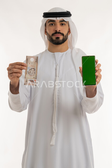 الأعمال الاقتصادية المالية، محاولة إدارة المصروف المنزلي بتدبر، بورتريه لرجل عربي خليجي اماراتي يرتدي الكندورة والغترة البيضاء ينظر الى الكاميرا بايماءات السعادة والسرور، عرض شاشة خضراء فارغة عبر الهاتف المحمول مع عملة الدرهم الاماراتي، خلفية بيضاء، كروما
